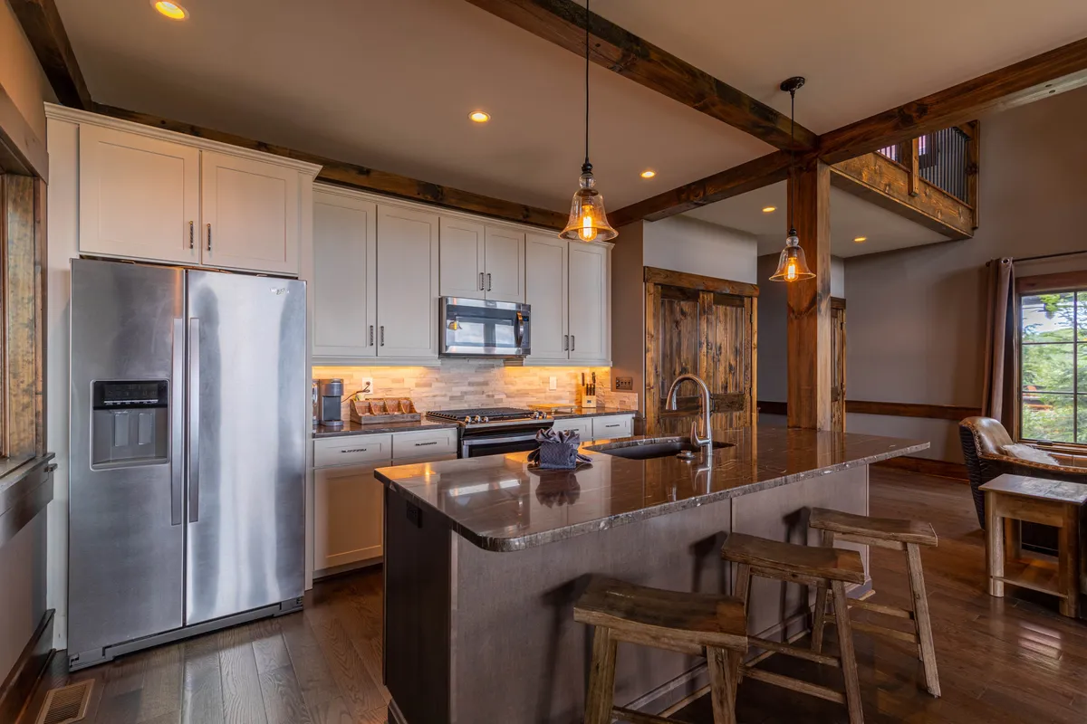 A modern kitchen with stainless steel appliances, a generous island countertop, wooden beams, and stylish pendant lighting. The space is bright and open, perfect for cooking and socializing, with a large window that hints at scenic views beyond.