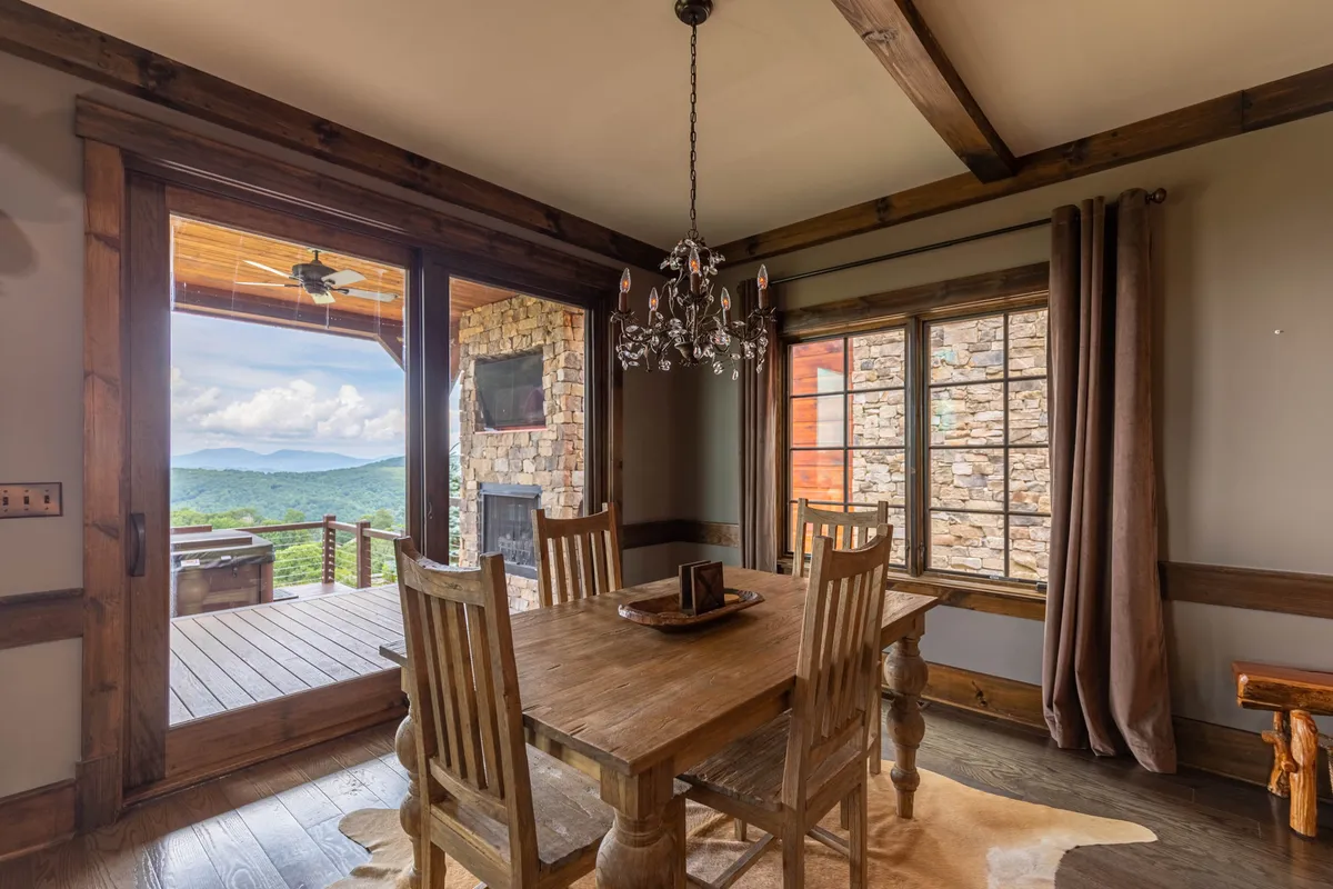 A cozy dining area featuring a wooden dining table, elegant chandelier, large windows, and a stone fireplace. The setting is bathed in natural light and offers panoramic mountain views, making it perfect for memorable family meals and celebrations.