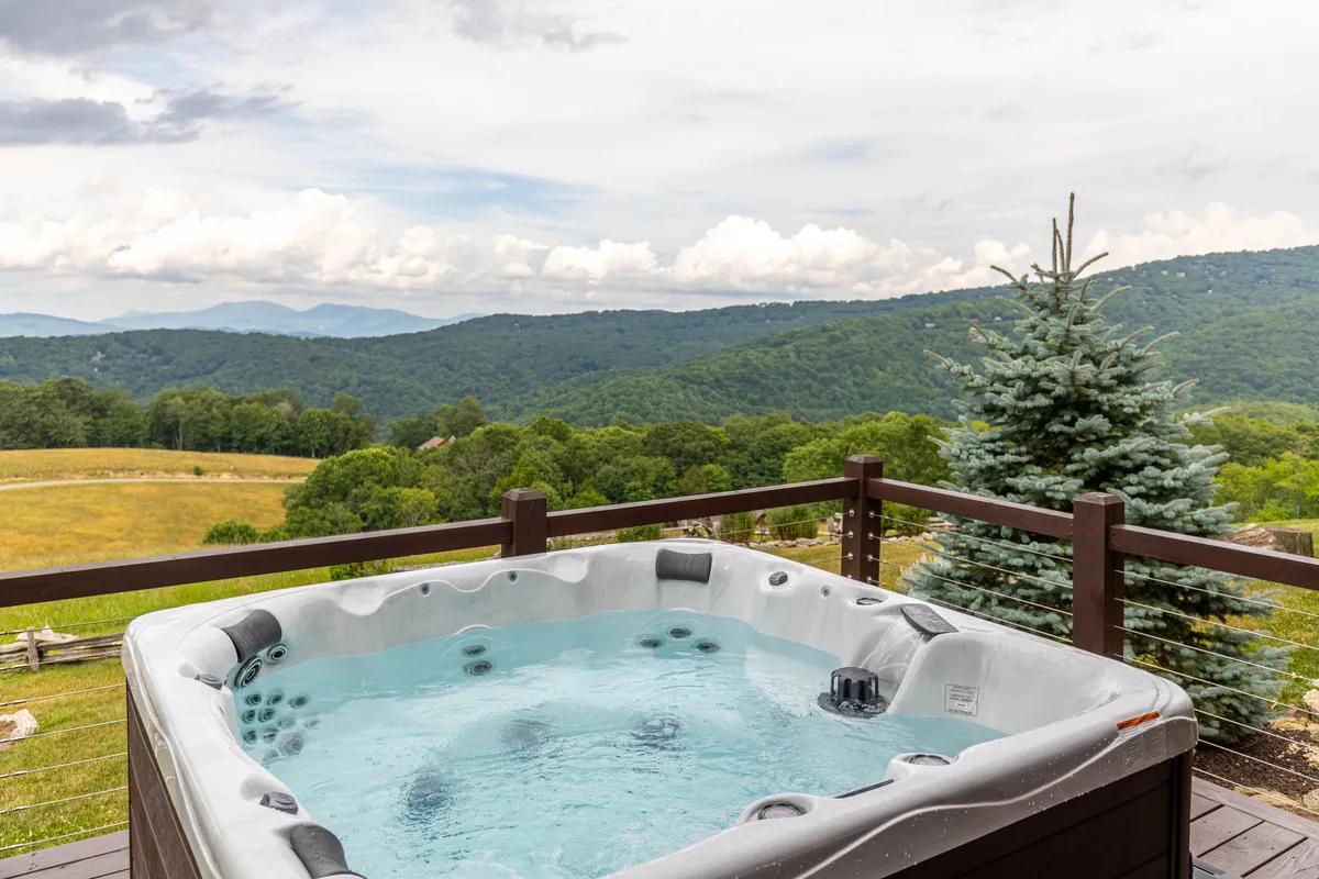 An outdoor hot tub sits on a deck that overlooks a breathtaking expanse of the Blue Ridge Mountains, surrounded by lush greenery. The scene radiates serenity, with the sky painted in sunset hues, inviting guests to unwind and soak in the views, perhaps with a loved one or close friends.