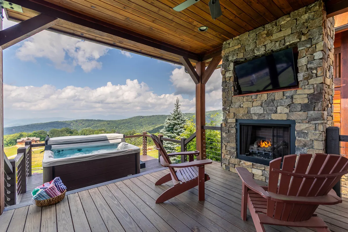 A spacious wooden deck featuring an inviting hot tub, cozy Adirondack chairs, a stone fireplace, and stunning mountain views, perfect for unwinding after a day of adventure.