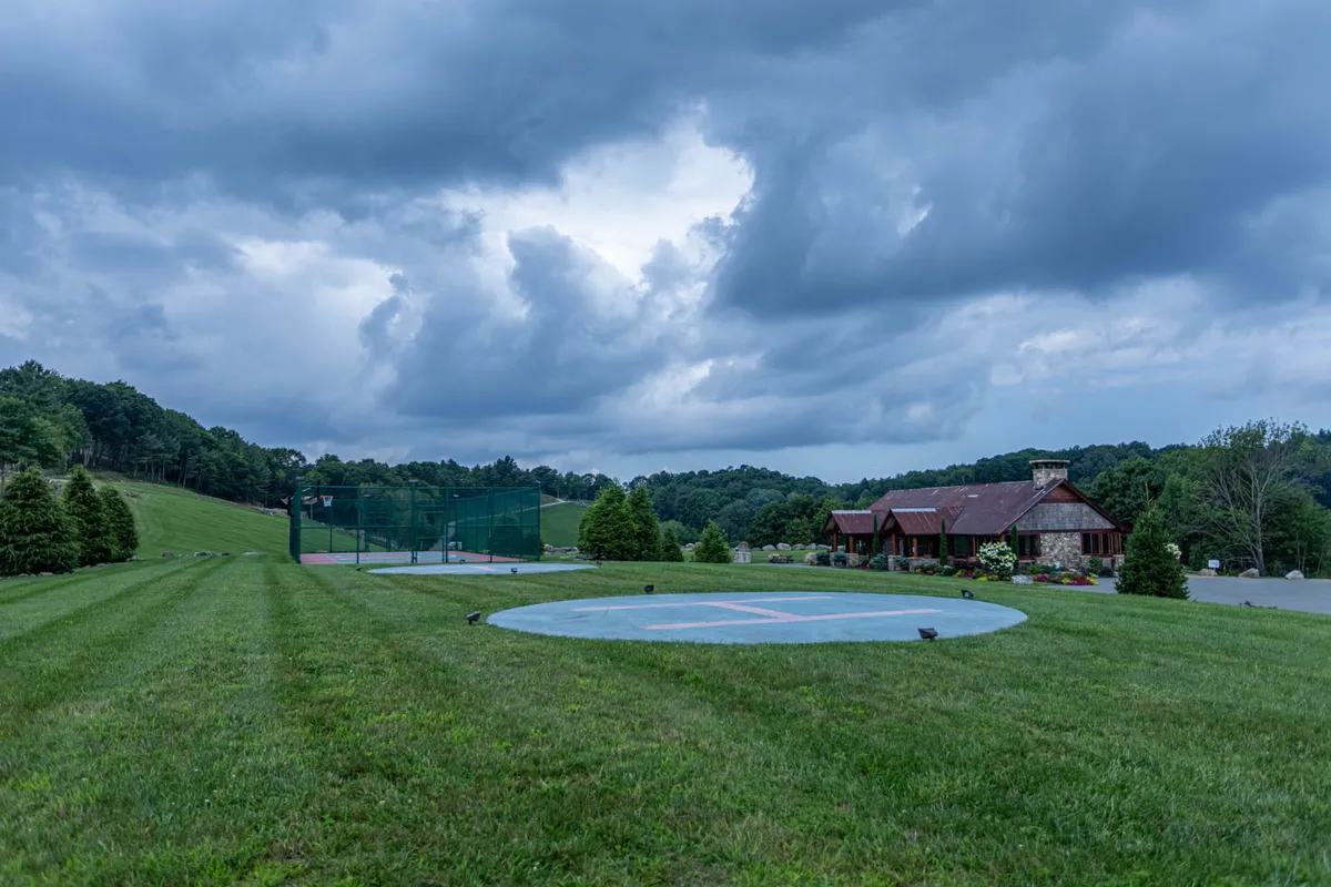 This exterior image shows a lush green lawn in front of the main lodge, with a helicopter landing pad visible and ample open space for outdoor gatherings. The area is set against a backdrop of trees and clear skies, giving a sense of space and community activity.