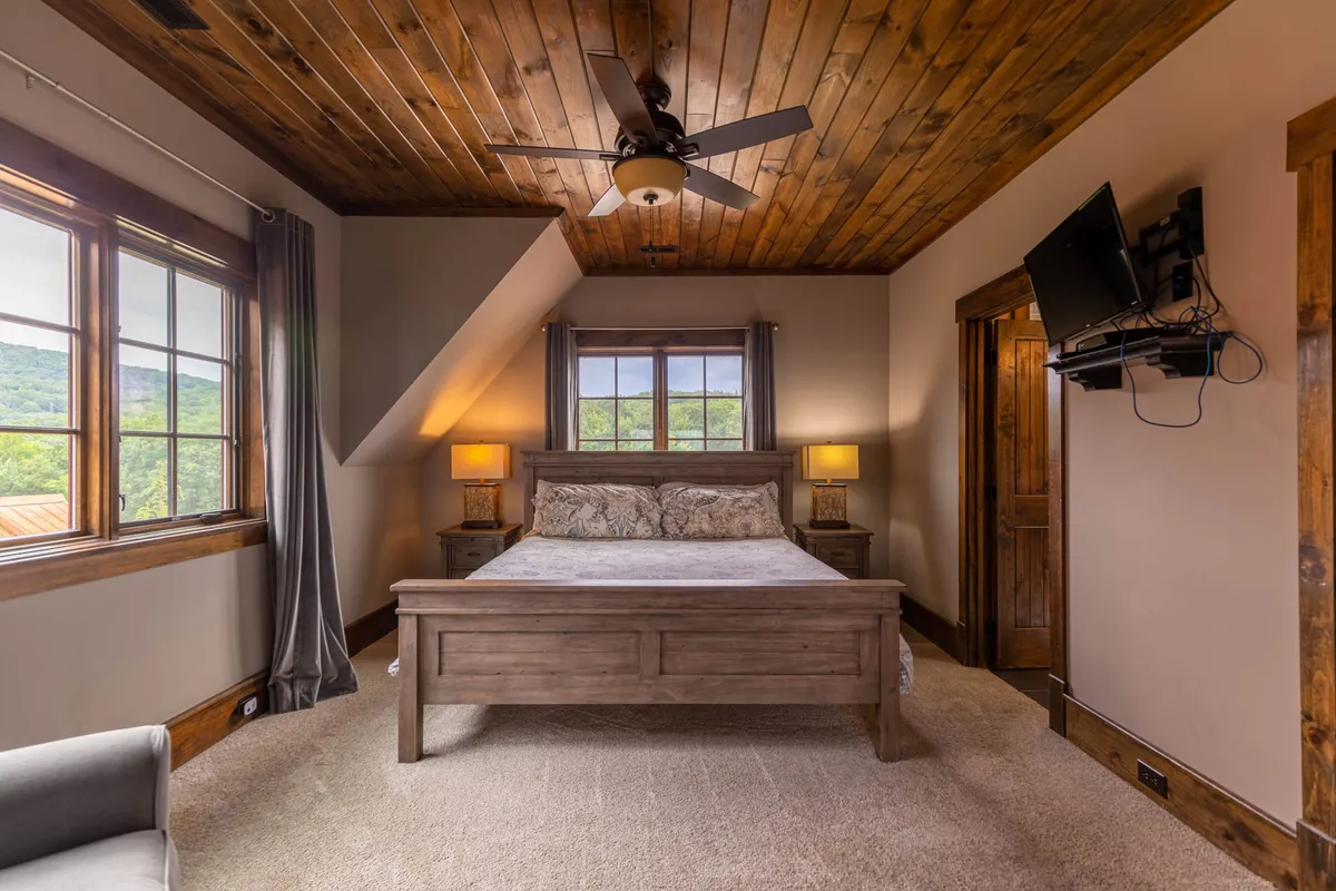 A spacious bedroom anchored by a plush king bed adorned with decorative pillows. Large windows allow abundant natural light to flood the room, highlighting the wooden ceiling and cozy nightstands with lamps. The atmosphere is tranquil and inviting, ideal for relaxation after a day outdoors.