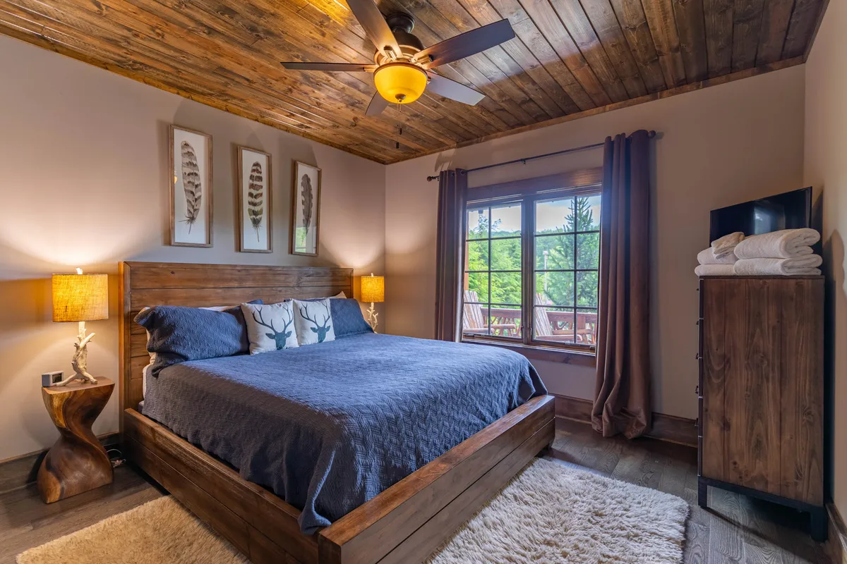 A calming bedroom setting features a wooden bed frame, decorative pillows, a window view, a nightstand with lamp, and a dresser with neatly stacked towels. The overall ambiance is serene and welcoming, perfect for restful nights or rejuvenating mornings.