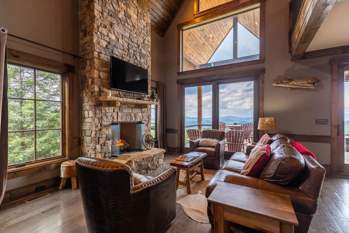 A spacious living room with a stone fireplace as the centerpiece, surrounded by comfortable seating. Large windows provide expansive views of the mountains and fill the room with natural light. The knotty-pine ceiling and wooden accents create a cozy, inviting atmosphere ideal for gatherings.