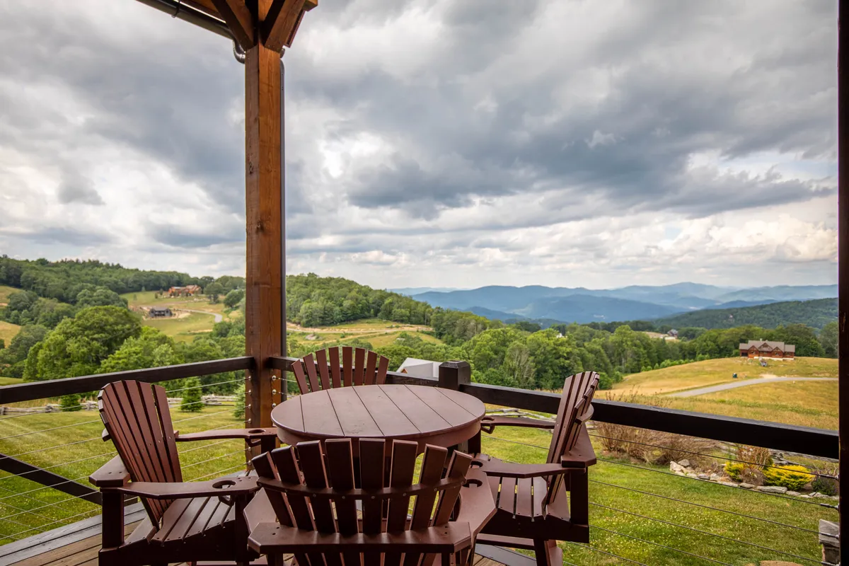 Two Adirondack chairs and a wooden table sit perfectly positioned to overlook rolling mountains under a tranquil sky, evoking a sense of peaceful seclusion and connection to nature. The setting suggests the ideal spot for morning coffee, sunset wine, or intimate conversation.