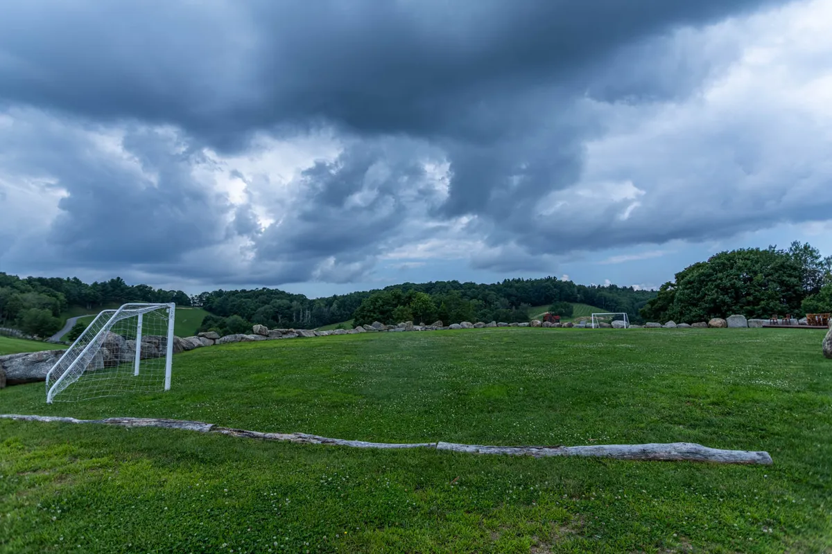 A wide, open green field with a soccer goal set against a backdrop of distant trees and a cloudy sky. The space provides plenty of room for outdoor play, sports, and picnics.