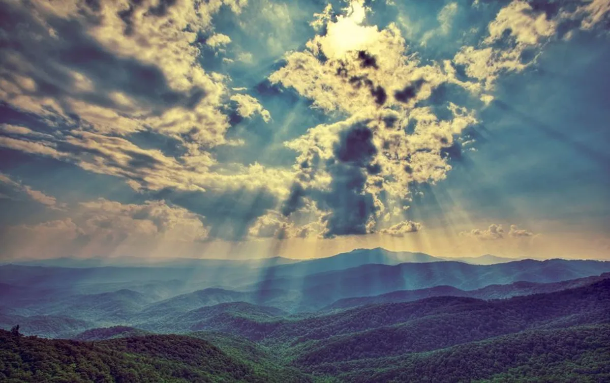 A wide landscape view of mountain ranges under dramatic cloud formations with rays of sunlight streaming through, inviting exploration. The setting inspires hiking, nature observation, and capturing the beauty of the natural surroundings.
